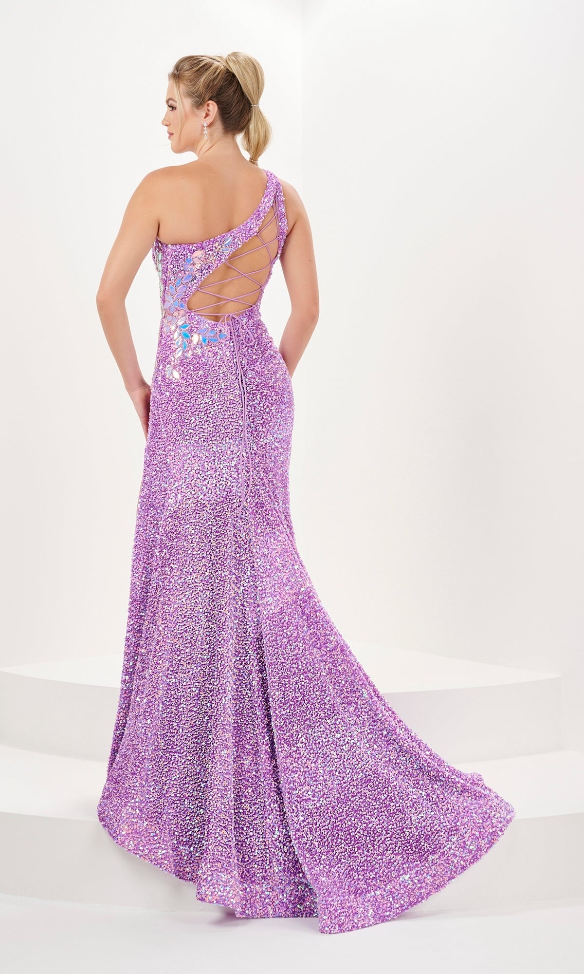 A woman stands on a white platform, facing away, wearing the Long Prom Dress 16061 by Tiffany—a floor-length, one-shoulder lavender velvet sequin dress with lace-up back and side slit.